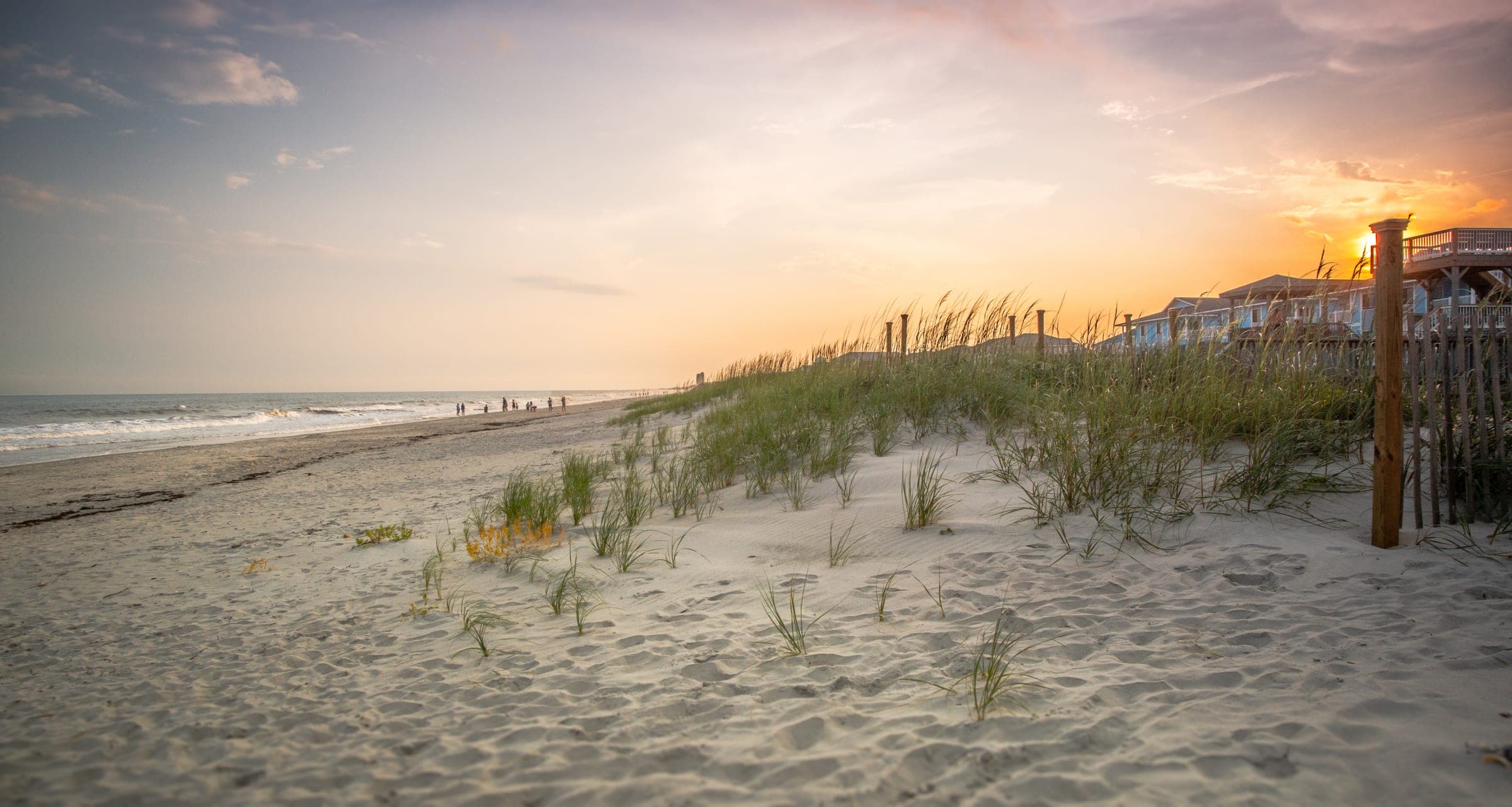 A wide Hamptons beach at sunset — white sand with footprints, dune grass and wildflowers in the foreground, beachgoers and ocean to the left, a row of shingled beach houses on the right, golden-hour sky