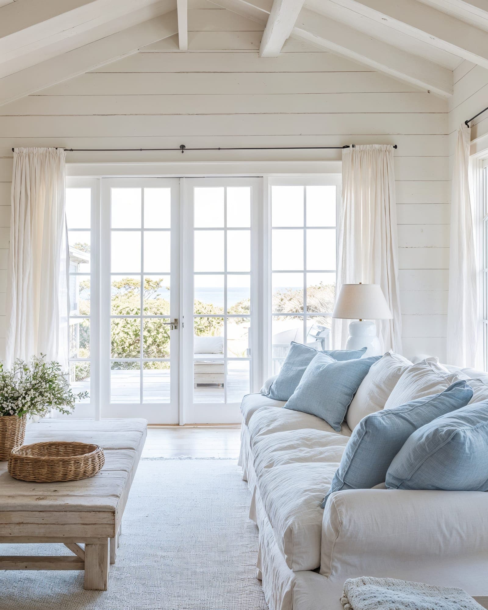 Sala de estar costera estilo Hamptons con sofá blanco, cojines azules y puertas francesas que dan al océano